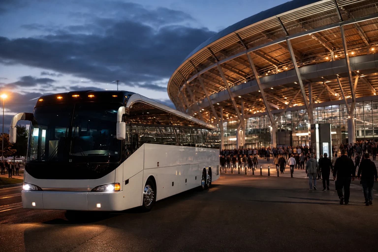 White coach parked outside a stadium at night as a crowd walks past